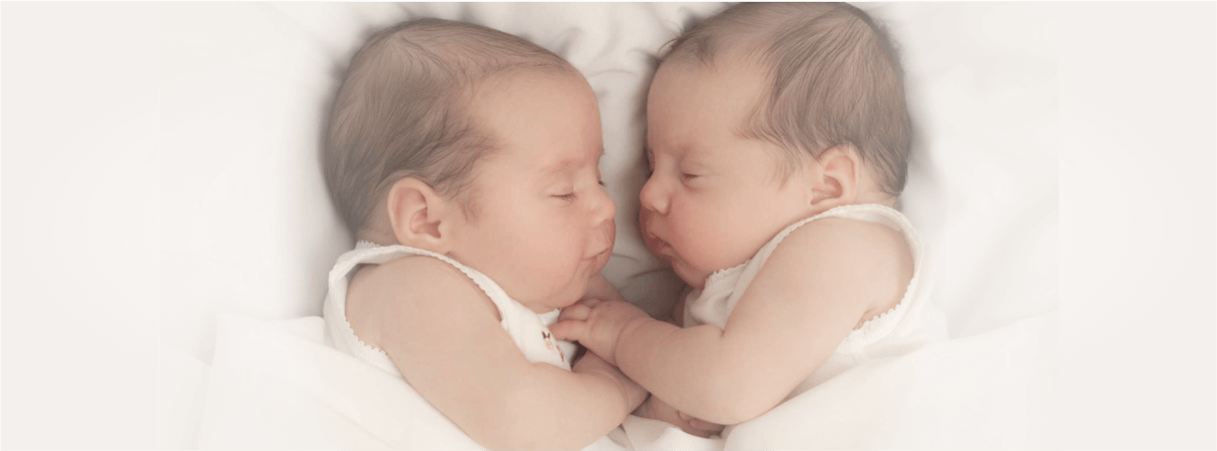 Two swaddled infants sleep facing each other on white blankets; soft lighting suggests serenity.
