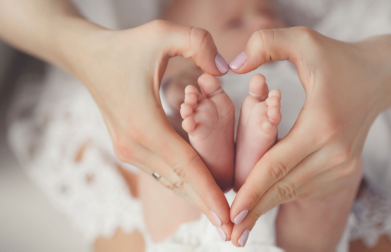 Hands form a heart around baby feet resting on white cloth; tender image of care and love.