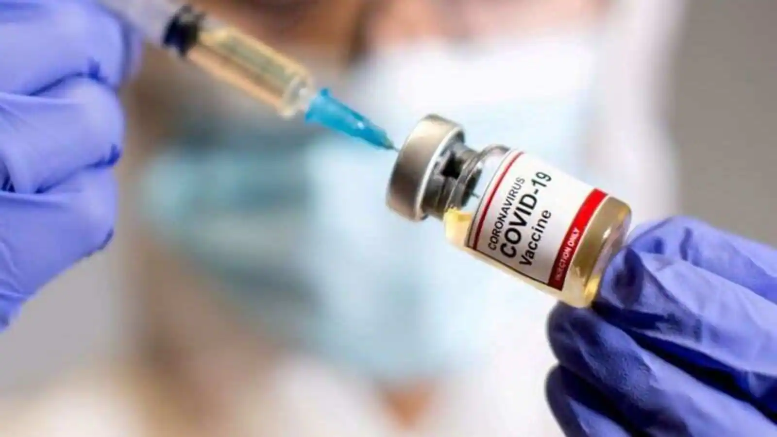 Gloved hand extracts COVID-19 vaccine from vial; background shows masked person in clinic.