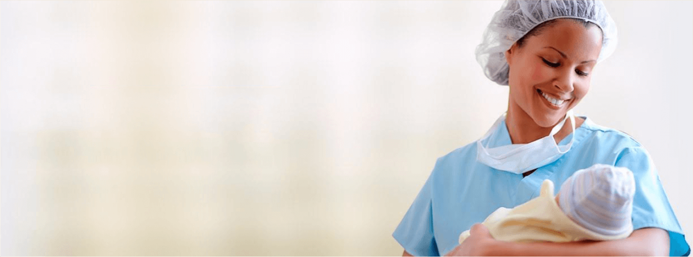 Nurse in blue scrubs cradles newborn in hospital setting; smiles warmly at the camera.